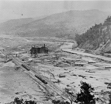Black and white photograph of Woodvale after a flood. A large brick building with chimneys is visible; it appears to be listing. There appears to be train tracks, train cars, and a few houses in what appears to be a plain of mud.