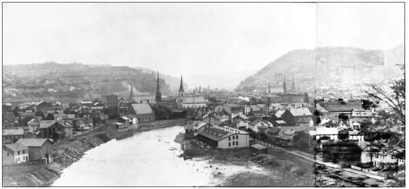 Black and white panorama of Johnstown before the 1889 flood. A stream with sharply angled but relatively shallow banks is in the foreground and curves to the right into the town. There appears to be some debris on the banks in the foreground, perhaps from earlier floods. 