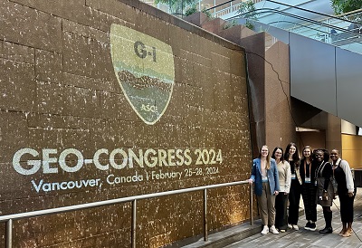 NC State Students smile next to a fountain. The G-I SHIELD and "Geo-Congress 2024" are projected on the fountain.