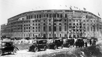Black and white photo of Yankee Stadium. In the foreground is a line of cars of the period, including a Model T