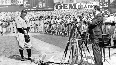 Famous black-and-white photograph of Lou Gehrig.  Gehrig stands at home plate in the left foreground, head bowed, cap in hand. At his feet is a trophy and some other objects. A man in a suit and a cluster of wooden tripods are in the right foreground. The Yankees are lined up behind them. In the background, Yankee Stadium is full.