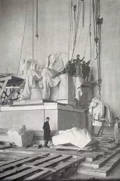 Assembly of statue of Abraham Lincoln. The statue is almost complete from its feet up to the shoulders. The left arm of the statue is being hoisted from the floor. 