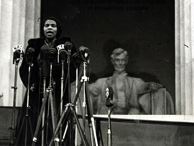 Marian Anderson sings on the steps of the Lincoln Memorial. The statue of Lincoln is visible in the background behind her left shoulder. She is wearing a hat and a dark fur coat, and is ringed by standing microphones.