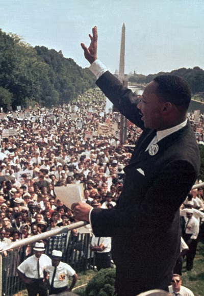 Color photograph of Martin Luther King addressing a crowd from the steps of the Lincoln Memorial. He is holding notes in his left hand; his right hand is raised as if he is waving to or greeting the crowd. The Washington Monument is in the background.