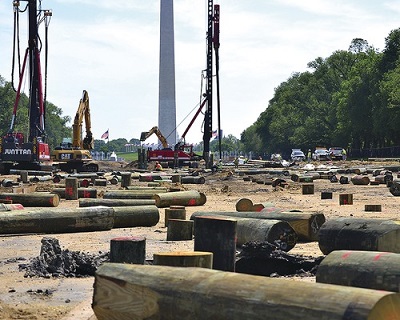 Color photo. Piles lie on the ground in the foreground; pile-driving equipment and excavators in the background; the Washington Monument in the far background.