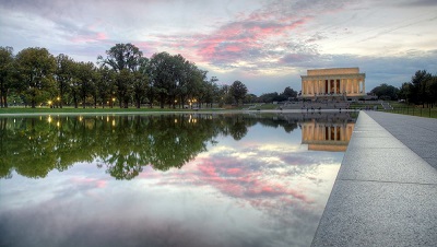 Color photograph of the Lincoln Memorial at twilight. The Memorial is lit and appears to be glowing, the clouds are pink, and the scene is mirrored in the Reflecting Pool.